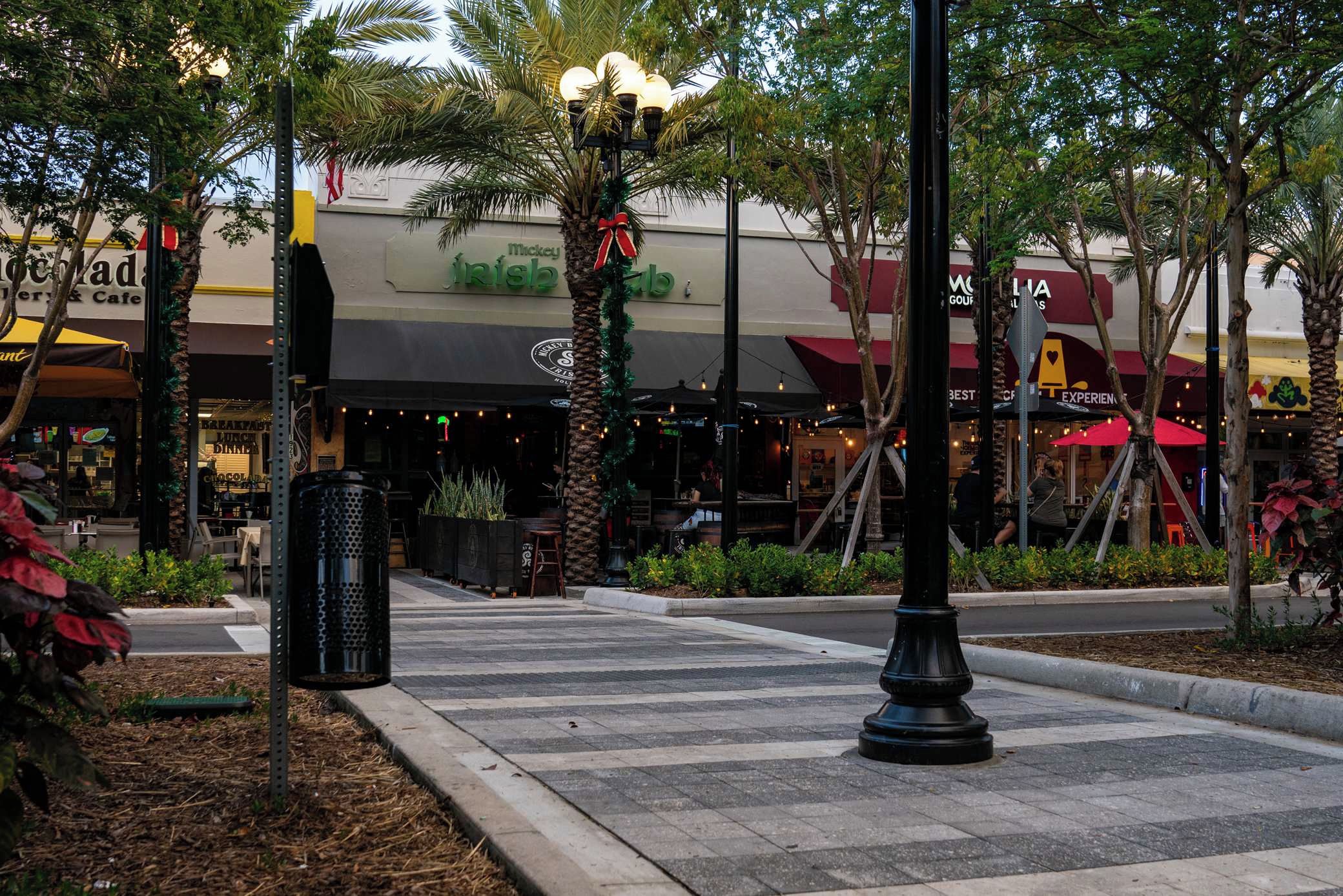 Zona peatonal con farol, palmeras y comercios, incluyendo un restaurante.
