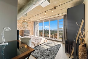 Een moderne slaapkamer met een groot bed, een bureau, een stoel, een raam met uitzicht op de stad en een bakstenen muur.