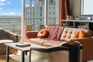 Una sala de estar moderna con un sofá, sillas, una mesa de centro y vistas a un paisaje urbano a través de ventanas amplias.