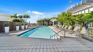 A swimming pool with lounge chairs and a covered seating area, surrounded by palm trees and a clear sky. - Midtown Rouge- Condo With City Views, Pool & Gym (Miami)