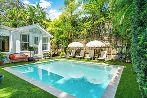 A swimming pool with lounge chairs and umbrellas, surrounded by lush greenery and a white house.