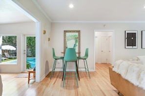 A room with a wooden floor, a bed with a white furry blanket, a dining table with chairs, and a painting on the wall.
