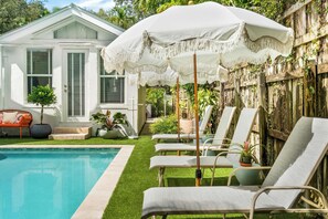 A pool area with lounge chairs, a white umbrella, and a house with shutters.