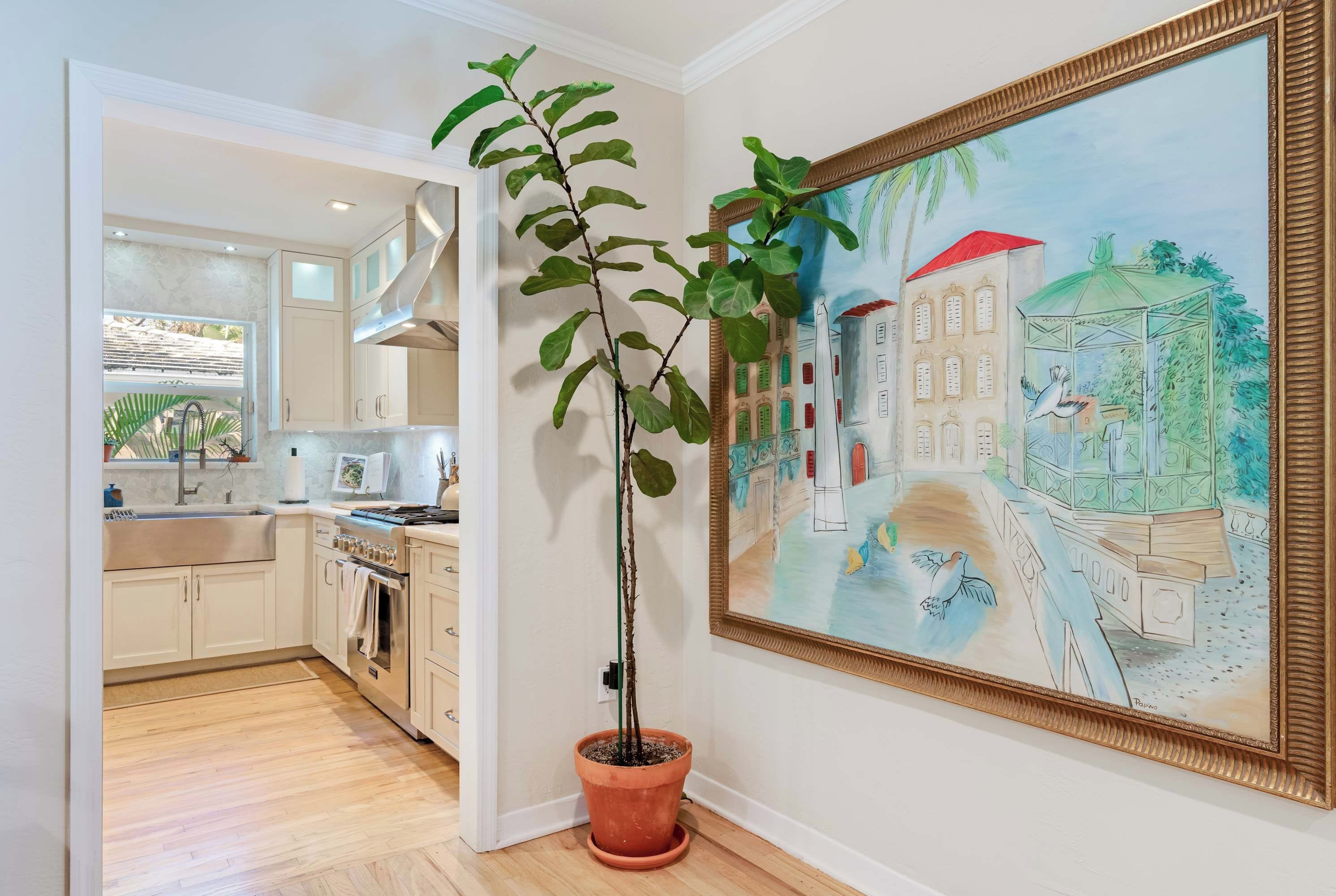 A kitchen with a potted plant, a painting, and a view into another room.