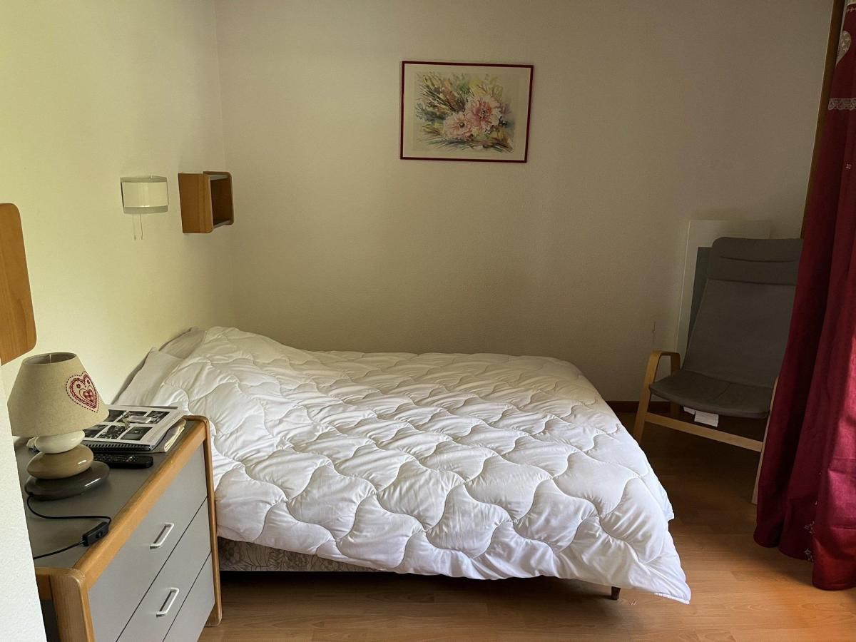 A single bed with a white duvet, a bedside table with a lamp, a framed picture on the wall, and a chair with a red curtain.