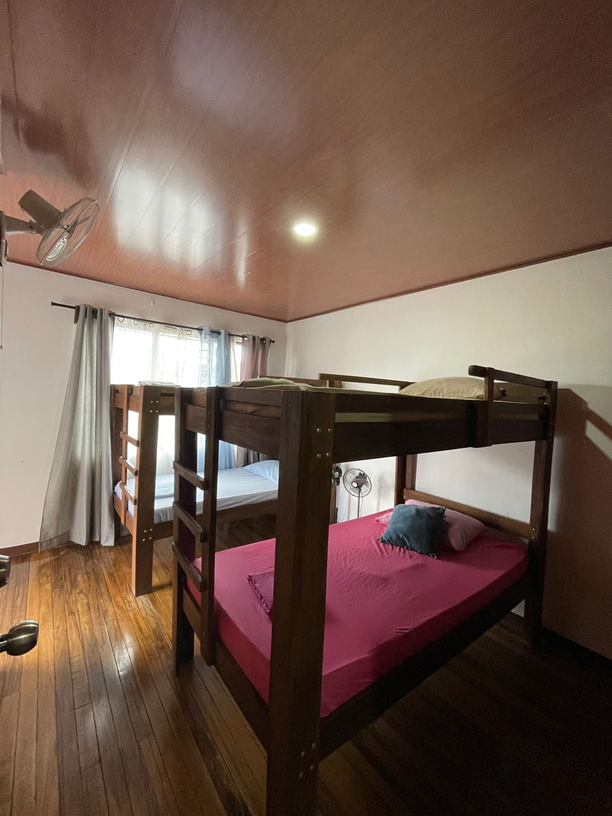 Dormitorio con literas, suelo de madera, ventilador de techo y ventana con cortinas.