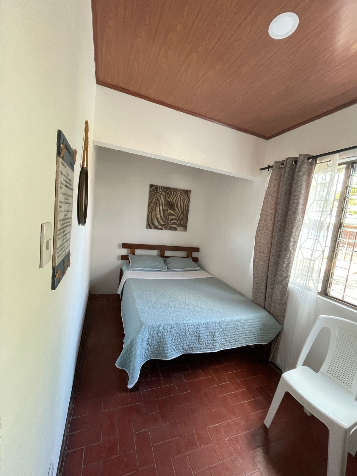 A bedroom with a bed, a headboard, a wall-mounted zebra artwork, a window with curtains, and a white plastic chair.
