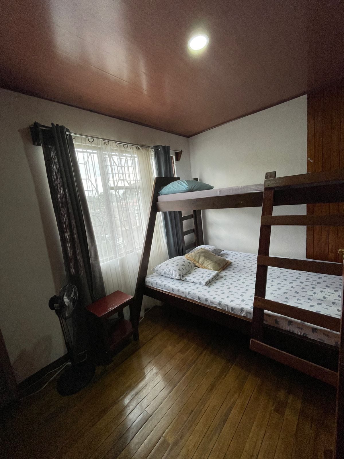 A bunk bed room with a wooden floor, a single bed with a pillow, a chair, a fan, and a window with curtains.