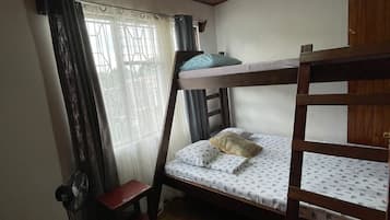 A bunk bed room with a wooden floor, a single bed with a pillow, a chair, a fan, and a window with curtains.