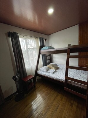 Habitación con literas, suelo de madera, una cama individual con almohada, una silla, un ventilador y una ventana con cortinas.