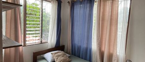 A single bed with a wooden frame, a red chair, a standing fan, and a window with metal bars.