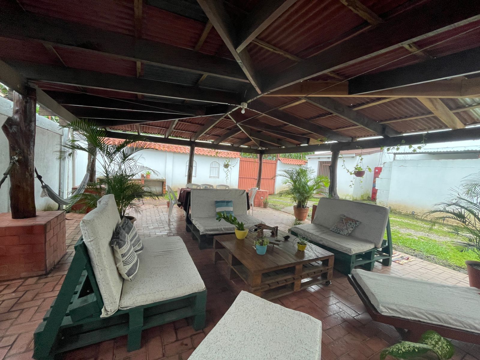 Un patio exterior con zona de estar techada, que incluye un sofá y un banco, rodeado de plantas en macetas.