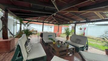 Un patio exterior con zona de estar techada, que incluye un sofá y un banco, rodeado de plantas en macetas.