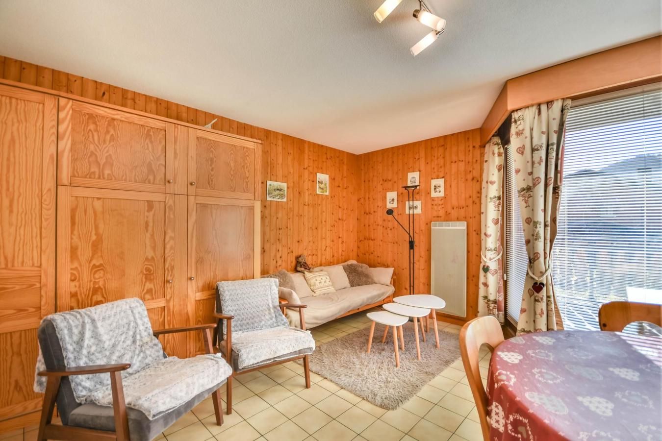 Um quarto aconchegante com paredes de madeira, um sofá, duas poltronas, uma mesa pequena e uma janela com cortinas.