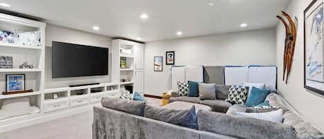 Una sala moderna con un televisor de pantalla plana grande, un sofá modular gris y un mueble de entretenimiento blanco.