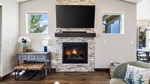 A living room with a stone fireplace, a television mounted above it, and a window with a tree view. - High West Hideaway by Avantstay Mountain-view Townhome Hot Tub + Firepit Near Park City (Heber City)