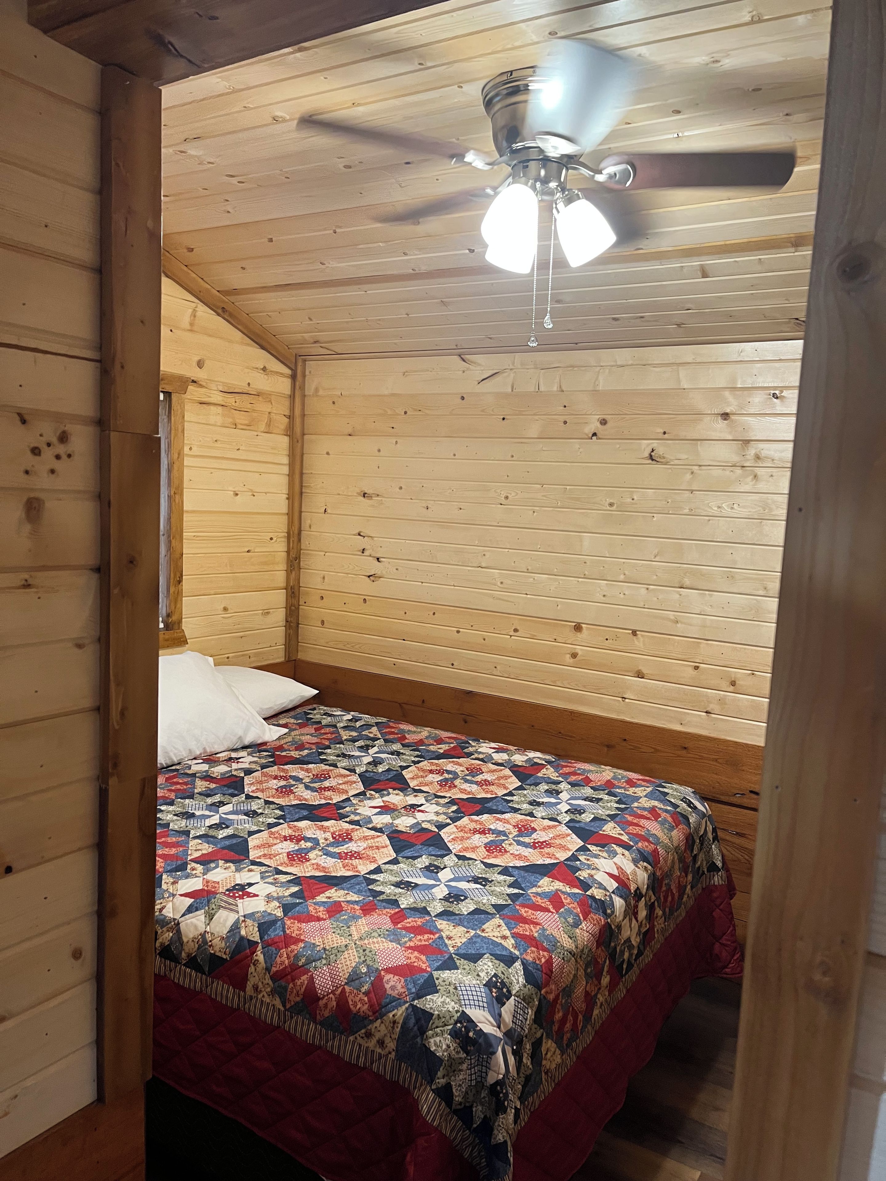 A cozy bedroom with a wooden ceiling and sloped roof, featuring a bed with a colorful quilt and a ceiling fan.