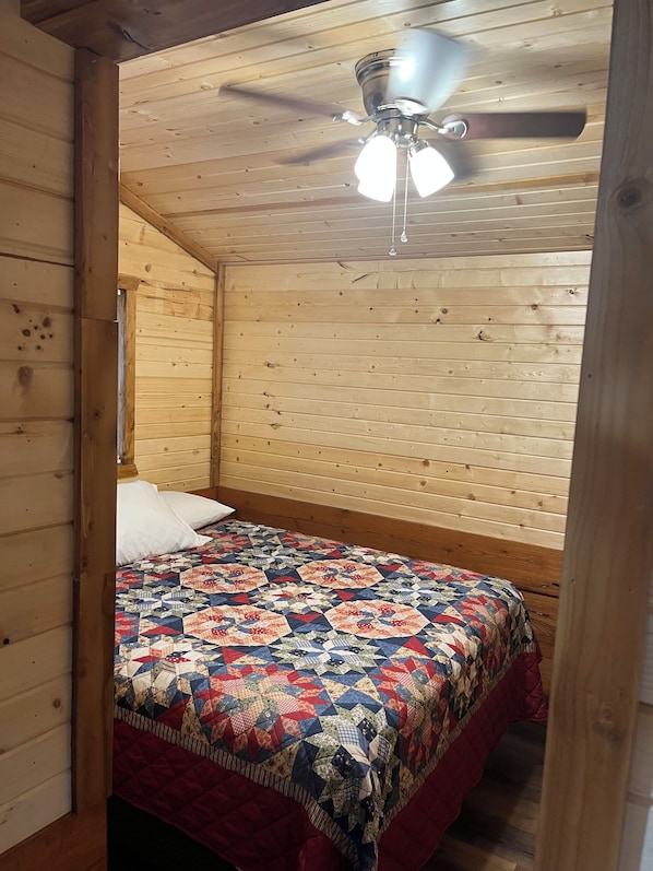 A cozy bedroom with a wooden ceiling and sloped roof, featuring a bed with a colorful quilt and a ceiling fan.