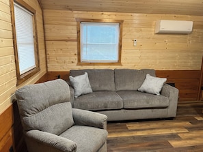 Ein gemütliches Zimmer mit grauem Sofa und Sessel, Holzwänden und einem Fenster mit Jalousien.