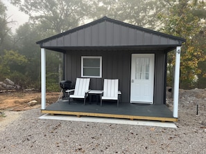 A small, dark-colored cabin with a porch, two Adirondack chairs, and a grill.