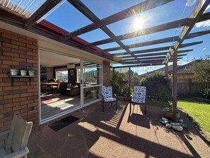 A covered patio area with a glass door leading to an interior space, a wooden pergola with hanging lights, and a brick wall. - Mooma's - Single Level, Covered Patio, Sleeps 8 (Rotorua)