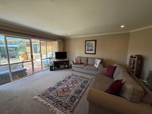 A living room with a sofa, a television on a stand, a patterned rug, and a large sliding glass door leading to an outdoor area. - Mooma's - Single Level, Covered Patio, Sleeps 8 (Rotorua)