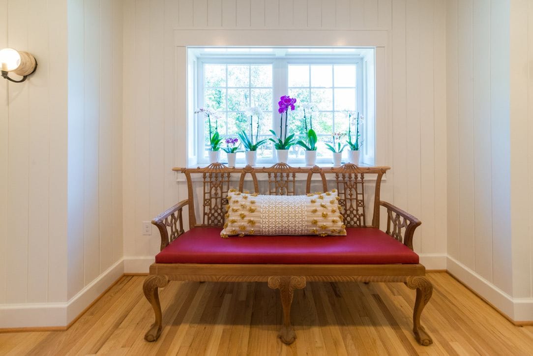 A wooden bench with a red cushion and a patterned pillow in front of a window with potted plants.