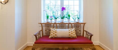 A wooden bench with a red cushion and a patterned pillow in front of a window with potted plants.