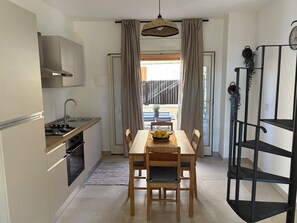 A compact kitchen with a dining area, a small table with chairs, a hanging light fixture, and a view of the outside. - Zora San Lorenzo - Marzamemi Beach (Reitani)
