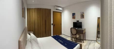 A hotel room with a bed, a desk, a chair, an air conditioning unit, and artwork on the wall.