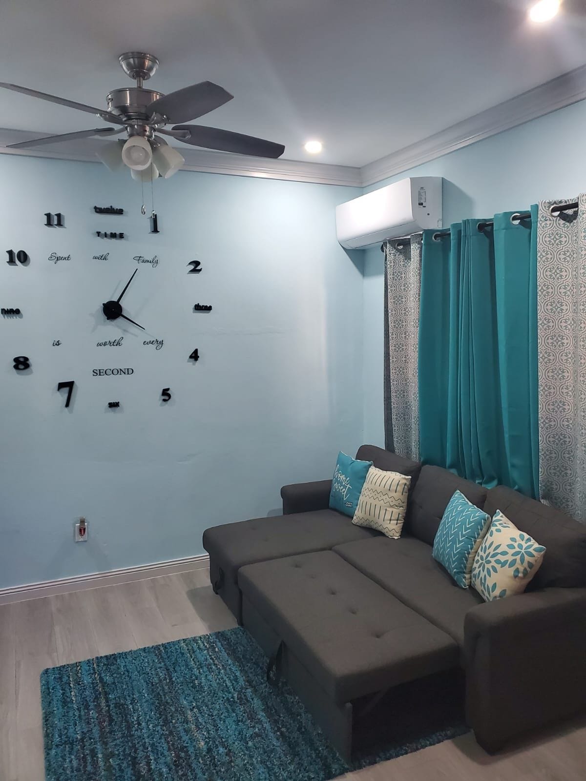 A living room with a sofa, a ceiling fan, teal curtains, and a clock on the wall.