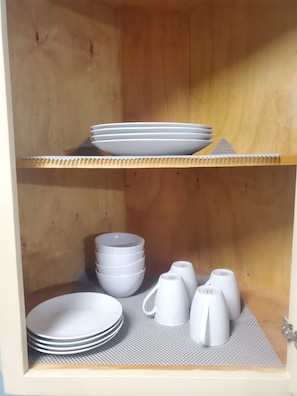 Ein Holzschrank mit Regalen, auf denen weiße Teller und Schüsseln stehen, und eine Ablage mit weißen Tassen und einer Teekanne.