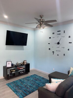 Ein Wohnzimmer mit Wandfernseher, Deckenventilator, Wanduhr, schwarzem Unterhaltungsschrank und blauem Teppich.