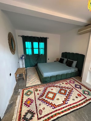 A bedroom with a bed, a small stool, a rug, a wall-mounted fan, and a window with curtains. - Private villa with a view of the lake—a peaceful retreat in Agafay. (Marrakech-Safi)