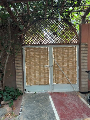 A gated entrance with a woven lattice roof, a metal gate, and a red and grey paved area leading to the gate.