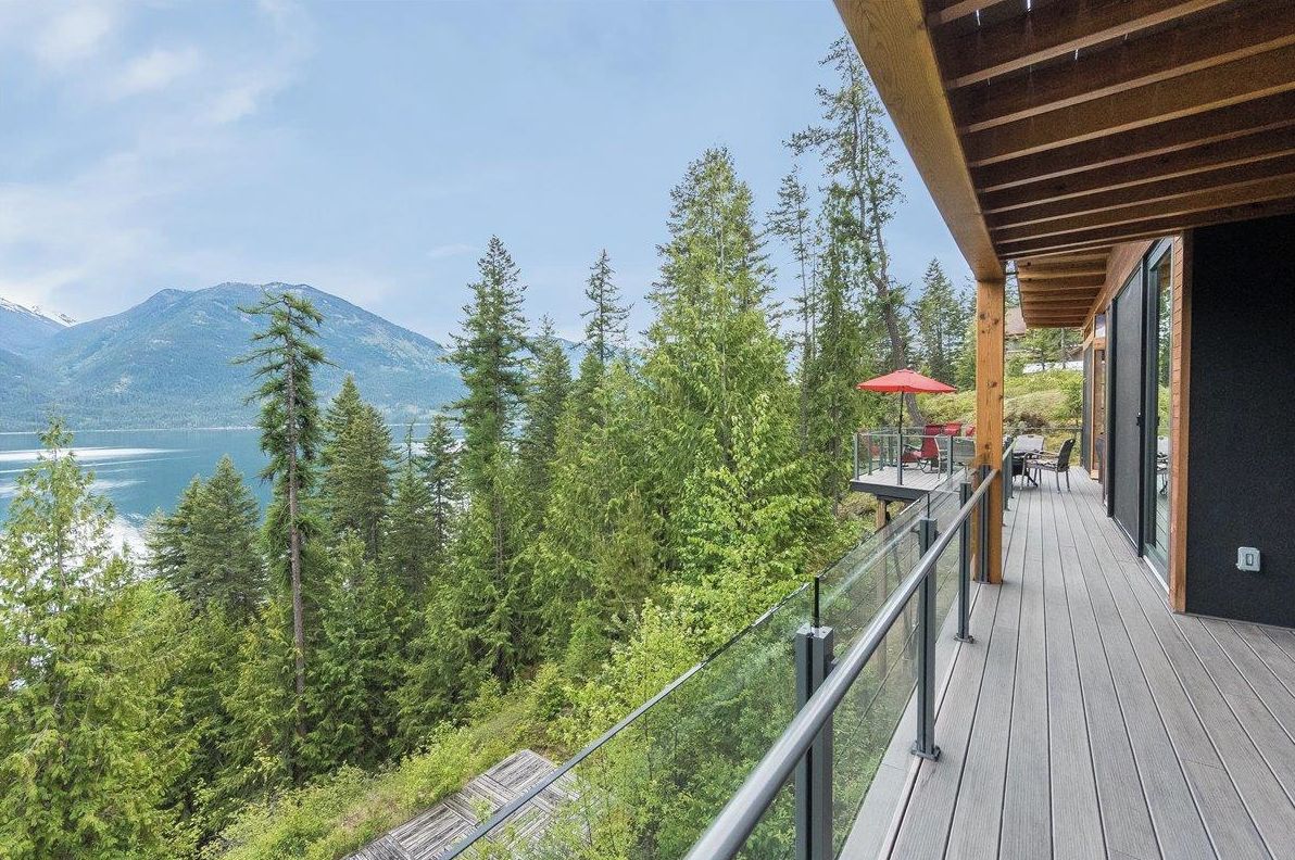 Eine Holzterrasse mit Glastrennwand, die auf einen See und bewaldete Berge blickt.