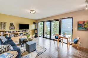 A living room with a sofa, coffee table, and a fireplace. There is a television, bookshelves, and a large window with a view.