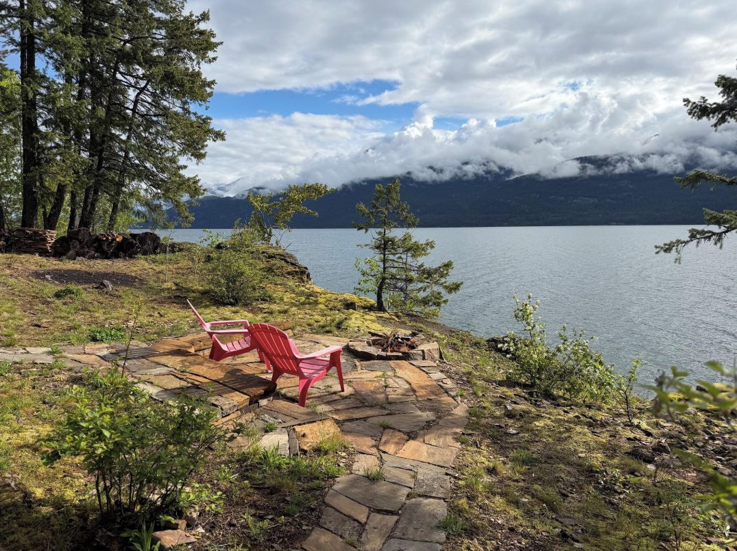 A lakeside setting with a red Adirondack chair on a stone deck, surrounded by trees and a view of a mountain range.