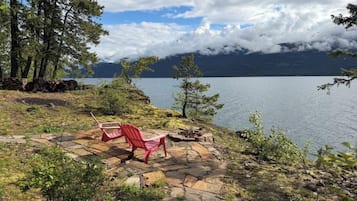 A lakeside setting with a red Adirondack chair on a stone deck, surrounded by trees and a view of a mountain range.