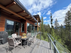 Um deck de madeira com mobiliário externo e guarda-corpo de vidro com vista para uma área arborizada.