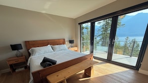 A bedroom with a wooden bed, a bench, a nightstand, a lamp, and a large sliding glass door leading to a balcony with a view of a lake and mountains.