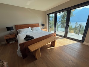 Quarto com cama de madeira, um banco, mesa de cabeceira, luminária e uma grande porta de vidro deslizante que dá para uma varanda com vista para um lago e montanhas.
