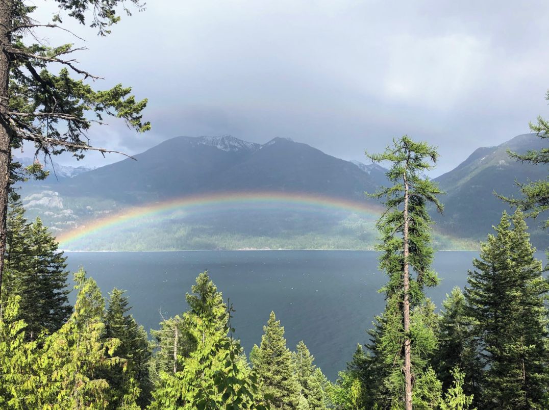 Ein See, umgeben von Bergen und Bäumen, mit einem Regenbogen.