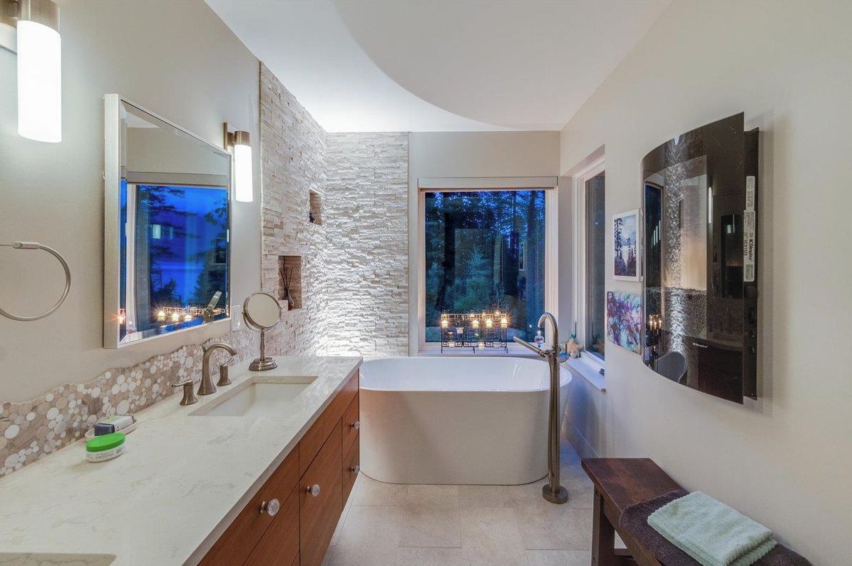 A modern bathroom with a freestanding bathtub, a large mirror, and a window with a view of trees.