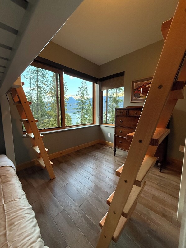 A room with a loft bed, wooden ladder, large windows, and a dresser.