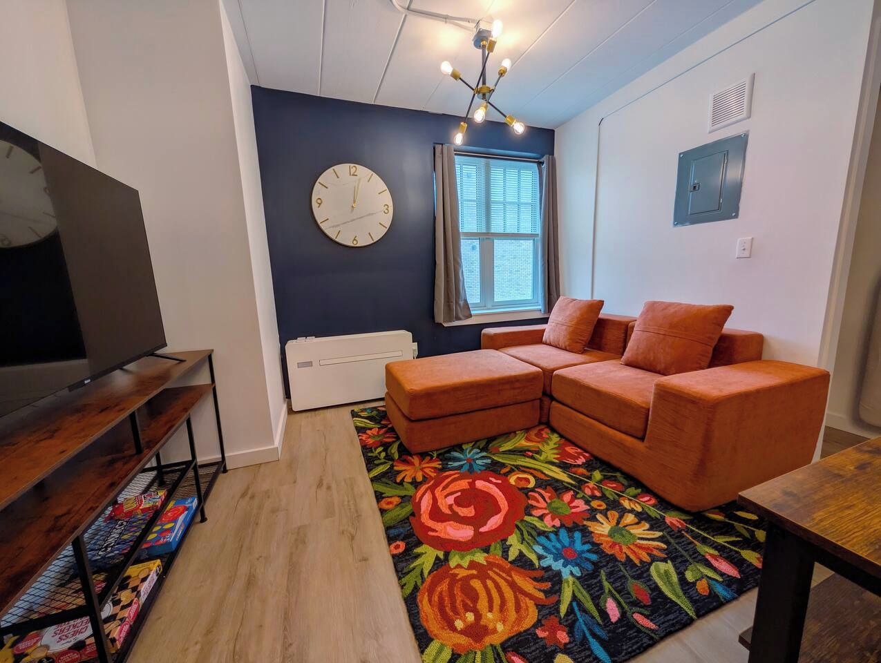 A modern living room with a sectional sofa, a flat-screen TV, a decorative rug, and a window with blinds.