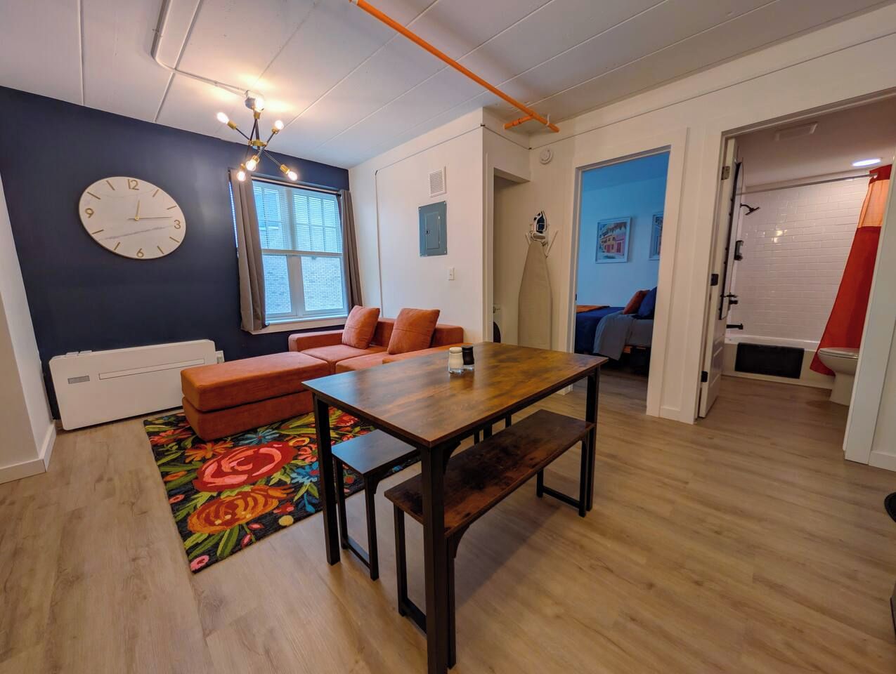 A room with a wooden dining table, a sofa, a clock, and a window.