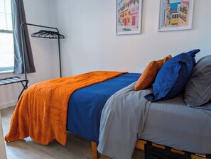 A bed with an orange blanket, a blue bedspread, and two pillows in a room with a window, a wall-mounted rack, and two framed pictures. - 1 Bdrm Gem: Walkable, Downtown, Queen Bed (Detroit)