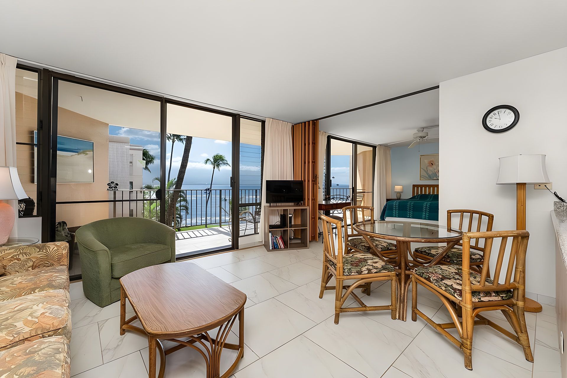 Una habitación de hotel con balcón, una zona de comedor con mobiliario de mimbre, un televisor y un reloj en la pared.