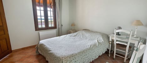 Un dormitorio con cama, una silla, una mesita de noche, una lámpara y una ventana con cortinas.